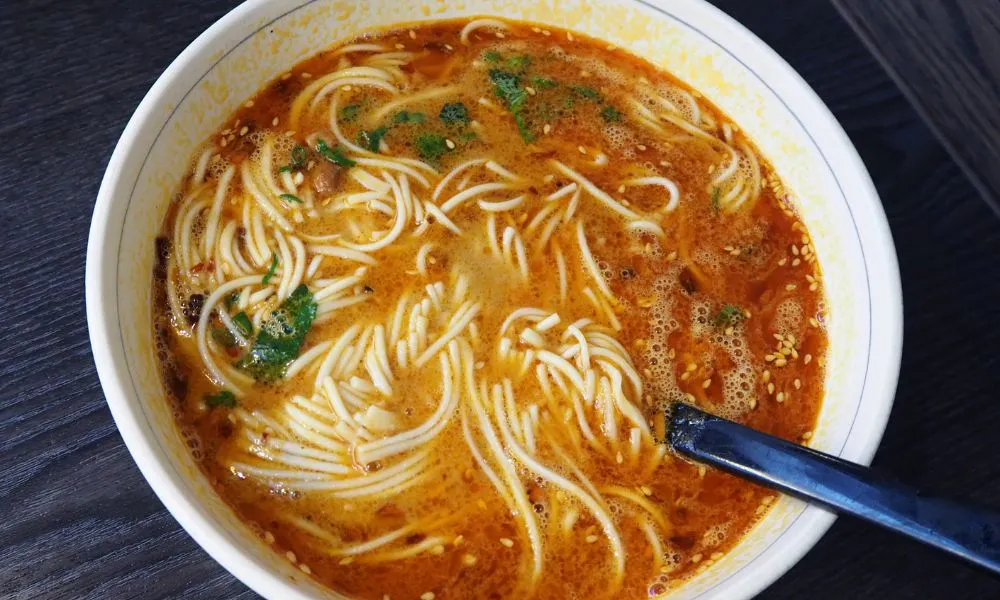 Spicy red broth noodle soup garnished with green herbs and sesame seeds at Good Alley, a Chinese restaurant in Arcadia
